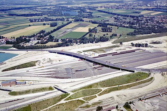 Der Rangierbahnhof in der Bauzeit 1990. Die neue Dachauer Straße auf dem langezogenen Brückenbauwerk überquert das Areal erst in westlicher Richtung, und schwenkt dann wieder in den Verlauf vor der Dachauer Straße in Karlsfeld ein. Hinter den Gleisanlagen und dem Wäldchen, die Ortschaft Ludwigsfeld mit der vormaligen Dachauer Straße, die ab 1992 Auf den Schrederwiesen genannt wird.